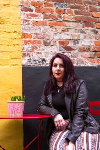 Lifestyle portrait of purple haired model sat down in front of a yellow wall